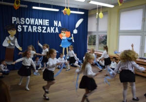 Na tle dekoracji dzieci stoją w półkolu, na pierwszym planie dziesięć dziewczynek tańczy układ taneczny z niebieskimi szarfami